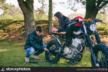 Young man fixing his damaged motorcycle while his girlfriend looks at him taking a beer outdoors. Man fixing his motorcycle while his girlfriend looks at him