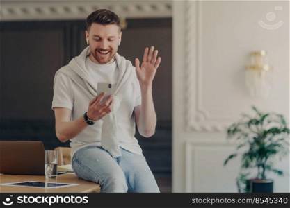 Young man freelancer looking at smartphone and greeting colleagues during web conference, home worker sitting on work desk wearing white tshirt and having sweater over his shoulders. Excited male freelancer having video call on mobile phone sitting on work desk