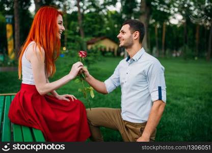 Young man gives flower to beautiful woman, romantic meeting of couple on a bench in summer park