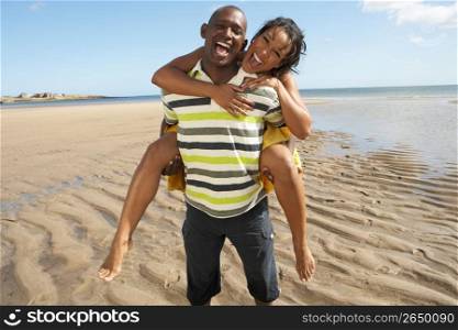 Young Man Giving Woman Piggyback Along Shoreline Of Beach