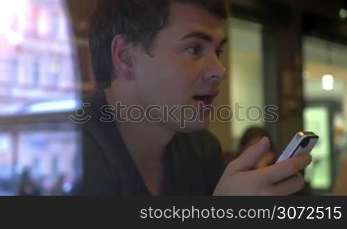 Young man having a lively conversation with somebody in a cafe while using smart phone. Outdoor view reflecting in the glass