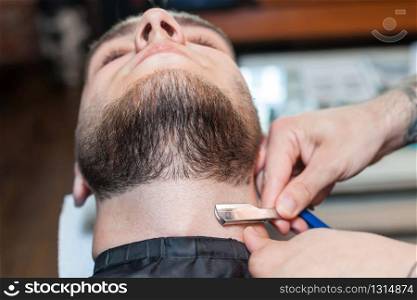Young man having his beard shaven, barber working with razor in the salon. Young man having beard shaven