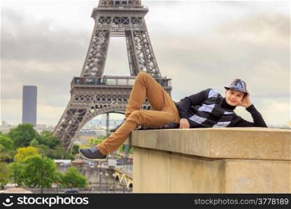 Young man hipster in a hat and vest lying on the parapet on the background of the Eiffel Tower (La Tour Eiffel) in Paris, France