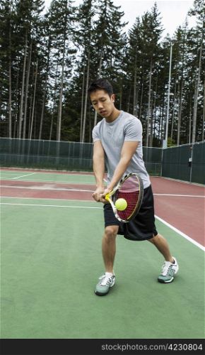 Young man hitting two handed backhand on outdoor tennis court with trees in background