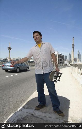 Young man holding a skateboard and hitchhiking at the roadside