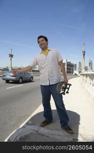 Young man holding a skateboard and hitchhiking at the roadside