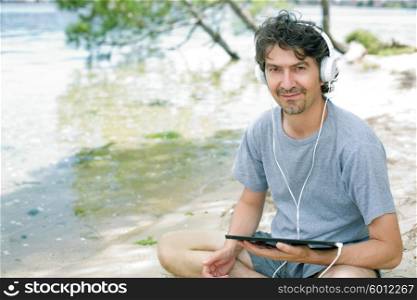 young man holding a tablet with headphones, at the beach