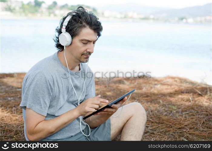 young man holding a tablet with headphones, at the beach