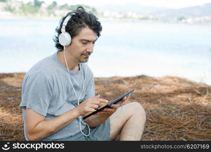 young man holding a tablet with headphones, at the beach