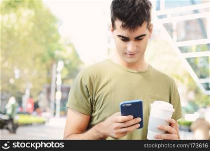 Young man holding coffee and reading message on his phone. Outdoors.