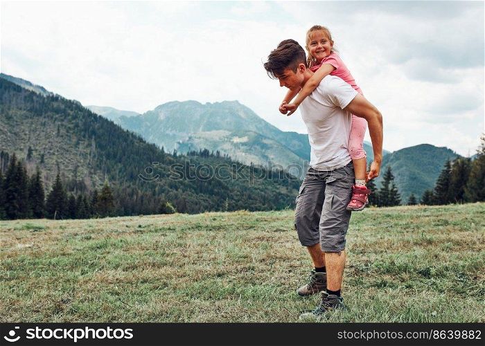 Young man holding little girl on his back. Child playing with her elder brother riding on his back enjoying summer day together. Happy siblings playing in the field during vacation trip in mountains. Young man holding little girl on his back. Child playing with her elder brother riding on his back enjoying summer day together