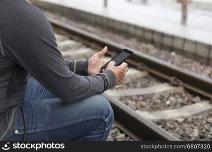 young man holding mobile phone sitting on platform at train station - travel concept