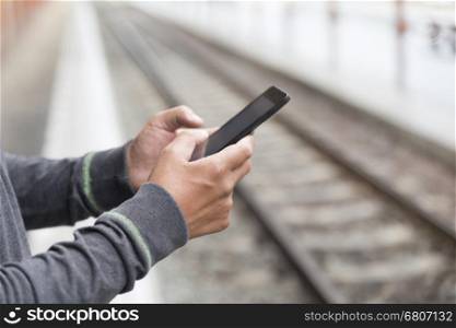 young man holding mobile phone standing on platform at train station - travel concept