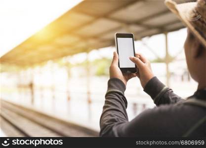 young man holding mobile phone with backpack standing on platform at train station - travel concept