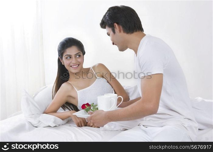 Young man holding tray of coffee cups and rose by woman in bed