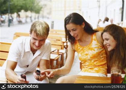 Young man holding two mobile phones with two young women sitting beside him