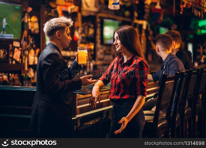 Young man holds glass of beer in hand and talks with woman at the bar counter in a sport pub, happy leisure of football fans