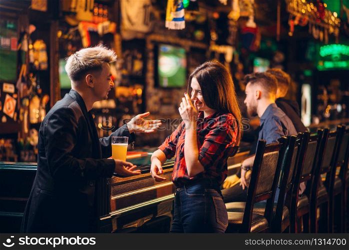 Young man holds glass of beer in hand and talks with woman at the bar counter in a sport pub, happy leisure of football fans. Young man talks with woman at the bar counter