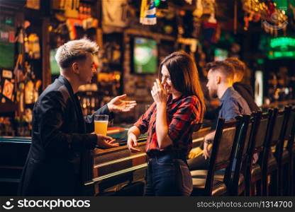 Young man holds glass of beer in hand and talks with woman at the bar counter in a sport pub, happy leisure of football fans. Young man talks with woman at the bar counter
