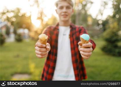 Young man holds out two ice creams in his hands in summer park. Youngster with ice-cream outdoors, male teenager with cold sweet