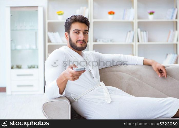 Young man in a bathrobe watching television at home on a sofa co. Young man in a bathrobe watching television at home on a sofa couch
