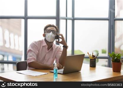 Young man in a face mask talking on the phone while working on his laptop
