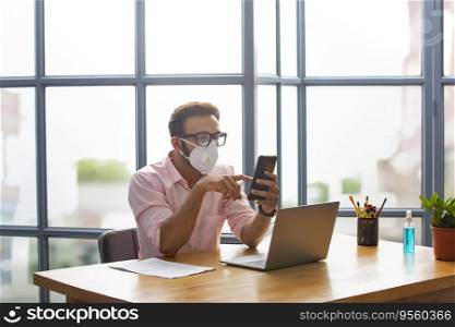 Young man in a face mask texting on the phone while working on his laptop