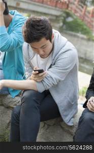 Young man in a gray hooded sweatshirt looking down at his phone and texting outdoors