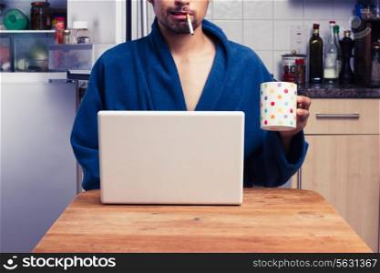 Young man in bathrobe drinking tea and smoking whilst working on laptop