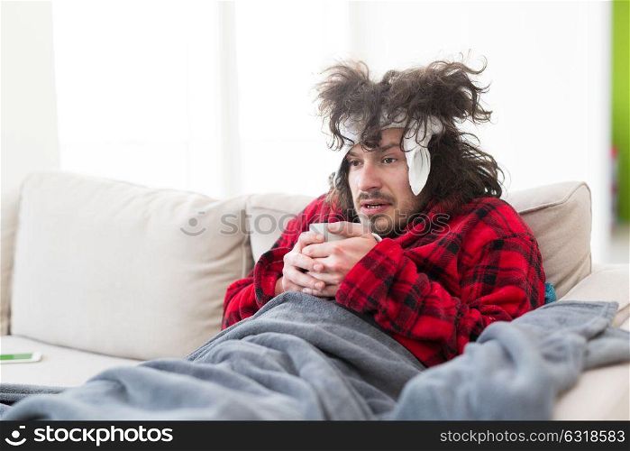 young Man in bathrobe with flu and fever wrapped holding cup of healing tea while sitting on sofa at home