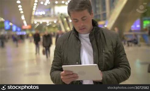 Young man in black jacket browsing the internet on tablet computer in crowded airport hall
