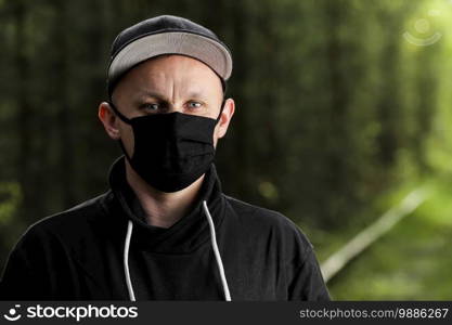 Young man in black protective antiviral mask in the summer park. The guy is resting outdoors on a sunny summer day in a mask made by his own hands. Virus protection. Quarantine measures.. Young man in black protective antiviral mask in the summer park. The guy is resting outdoors on a sunny summer day in a mask made by his own hands. Virus protection. Quarantine measures
