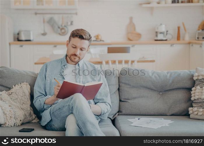Young man in casual clothes sitting on couch with legs crossed, writing down information in red note book, taking important notes with pencil, organizing and planning his day. Young man writing in note book while sitting on couch