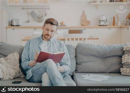 Young man in casual clothes sitting on couch with legs crossed, writing down information in red note book, taking important notes with pencil, organizing and planning his day. Young man writing in note book while sitting on couch