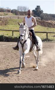 Young man in casual outfit riding white horse on sandy ground