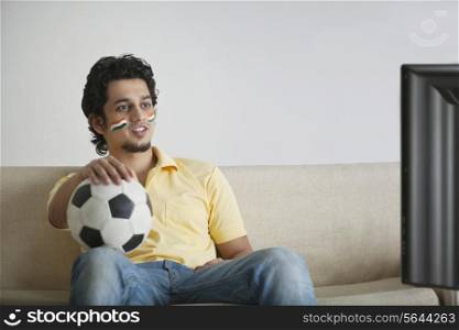 Young man in casuals with face painted watching television while holding soccer ball