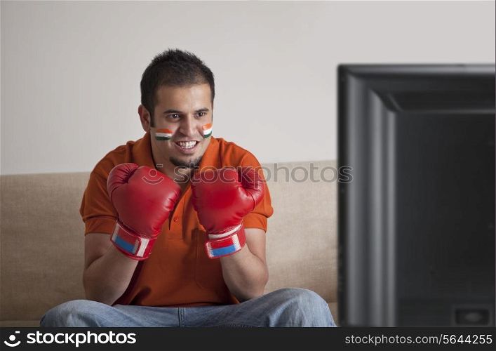 Young man in casuals with face painted wearing boxing gloves watching television at home