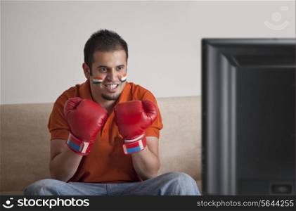 Young man in casuals with face painted wearing boxing gloves watching television at home