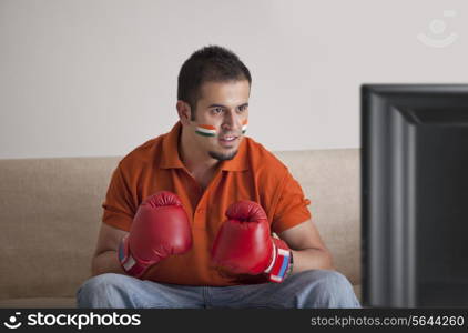 Young man in casuals with painted face wearing boxing gloves watching television at home