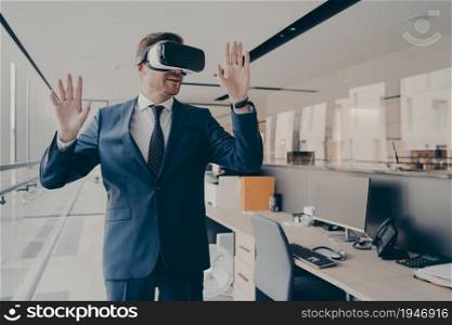Young man in formal suit enjoying virtual tour or excursion with VR headset. Businessman in virtual reality glasses standing near working desk in office, tries to touch objects in digital actuality. Businessman in virtual reality glasses standing near working desk in office