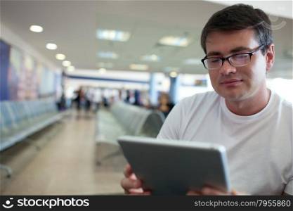 Young man in glasses using touch pad for reading or watching films while waiting at the airport or station