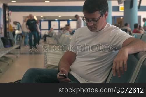 Young man in glasses with cell phone in the waiting-room of the airport or station. He browsing the internet on mobile to pass the time