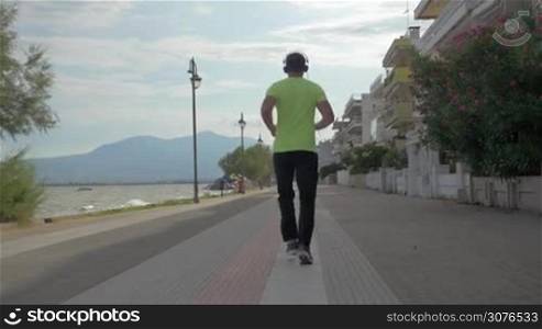 Young man in headphones runs on road of city Perea, Greece. On left side seen the sea, on right side homes