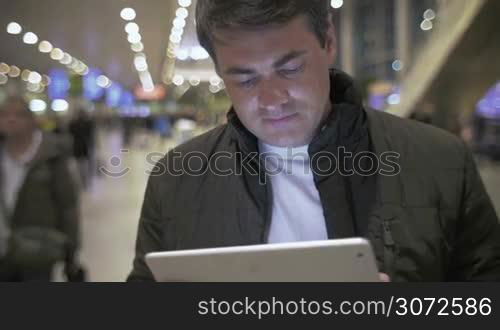 Young man in jacket using tablet computer at the crowded airport. Easy work with portable device