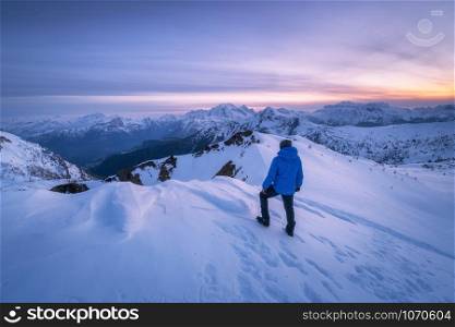 Young man in snowy mountains at sunset in winter. Sporty guy on the mountain peak, snow covered rocks and colorful blue sky with clouds at dusk. Travel in Dolomites. Wintry landscape with tourist