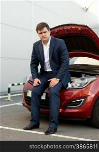 Young man in suit sitting on broken car with open hood