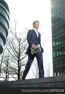 Young man in suit walking on wall outside office building holding newspaper