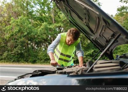 Young man in the reflective vest at the opened hood, car breakdown. Broken automobile or repairing of vehicle, trouble with auto engine on highway