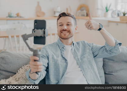 Young man in video call expressing his satisfaction, holding gimbal with phone attached, making thumbs up gesture and showing it to camera while sitting on couch, blurred background. Young man in video call expressing his satisfaction by showing thumbs up