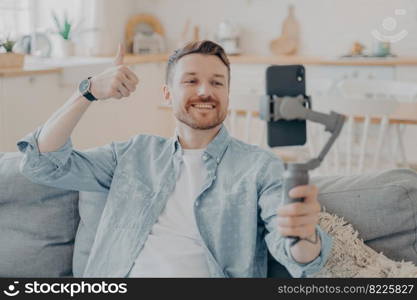 Young man in video call expressing his satisfaction, holding gimbal with phone attached, making thumbs up gesture and showing it to camera while sitting on couch, blurred background. Young man in video call expressing his satisfaction by showing thumbs up